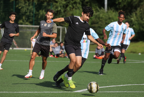 Zu sehen sind zwei Teams, die gegeneinander Fußball spielen. Ein Spieler läuft mit dem Ball auf die Kamera zu. Eine Mannschaft trägt Trikots mit der Aufschrift